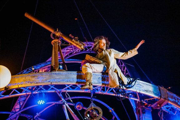 comédiens théâtre de rue Un personnage suspendu sur une structure métallique, pointant vers le ciel avec un télescope.