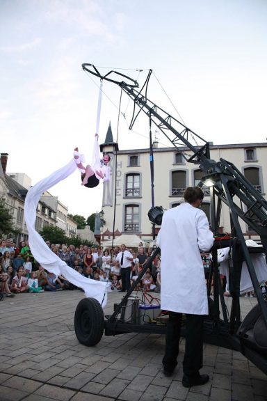 déambulation original Un artiste suspendu, vêtu de blanc, performe avec une structure métallique sur une place.