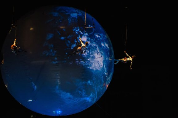 vertical dancing Performers are suspended near a glowing, stylized globe in a dark setting.