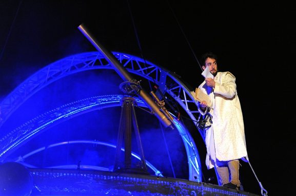 comédien galileo Un homme en manteau blanc souligne un canon sous des lumières bleues.