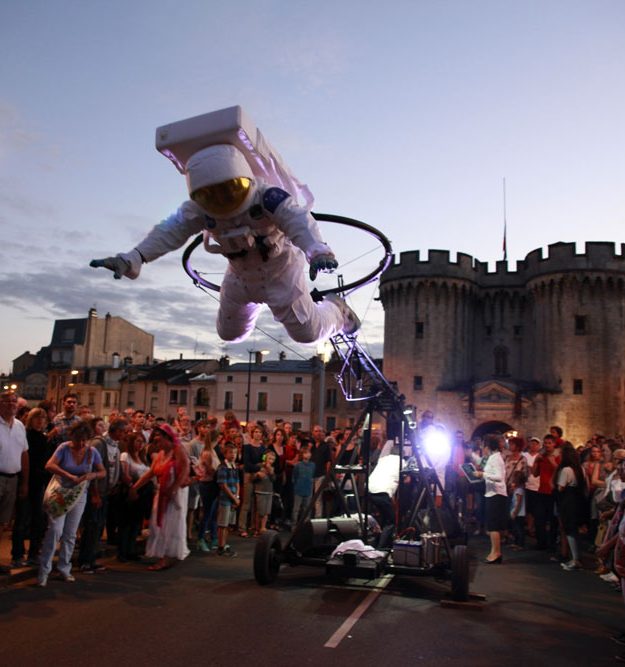 spectacle deambulation allégorie de la grenouille Astronaute volant au-dessus d'une foule, avec un fond de bâtiments historiques.