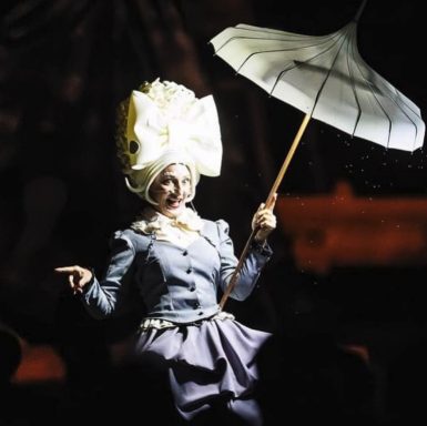 artiste de cirque Personne portant un chapeau extravagant, tenant un parapluie, sur scène dans une ambiance sombre.