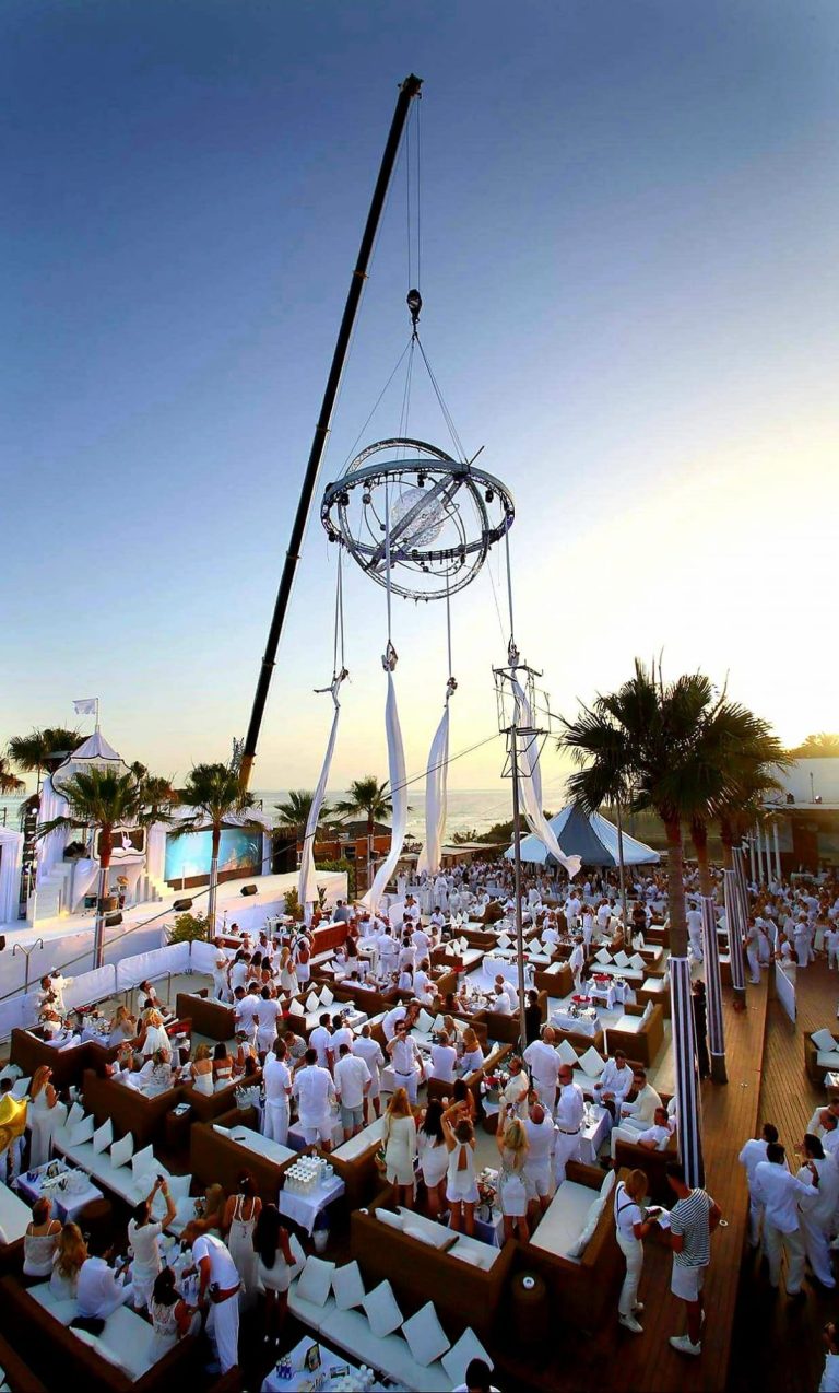 aerial show nikki beach club Marbella Événement en plein air avec des tables en blanc, une grue et un ciel bleu au crépuscule.