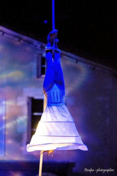 artiste a la corde lisse Une personne suspendue à un fil, vêtue d'une robe blanche et bleue.