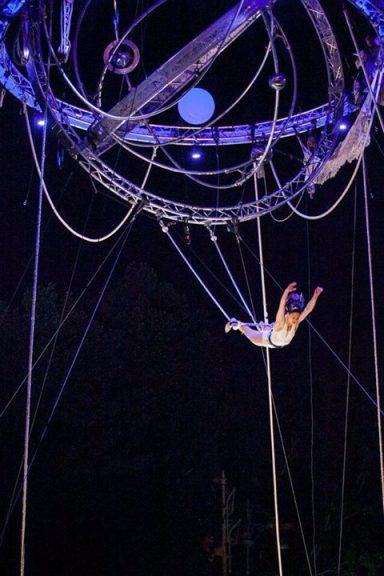 trapèze Cirue Artiste de cirque suspendu dans les airs, entouré de cercles métalliques et de câbles.
