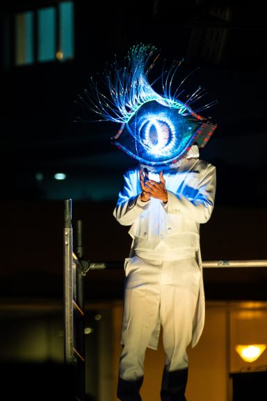 comédien de rue Personne en costume blanc tenant un symbole lumineux en forme d'œil sur scène.