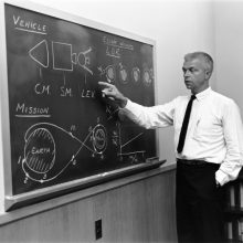 John le technicien de la nasa Un homme en costume montre un tableau noir avec des schémas de vaisseaux spatiaux.