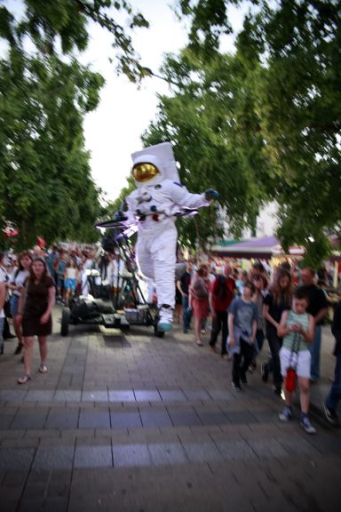 spectacle déambulation Un personnage géant en costume d'astronaute défile devant une foule sur une rue animée.