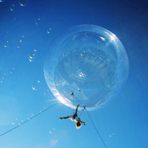 spectacle Bulle Une artistye suspendu à une bulle géante dans un ciel bleu parsemé de bulles.