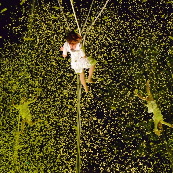 acrobate corde volante Une personne en robe blanche suspendue dans les airs, entourée de confettis verts.