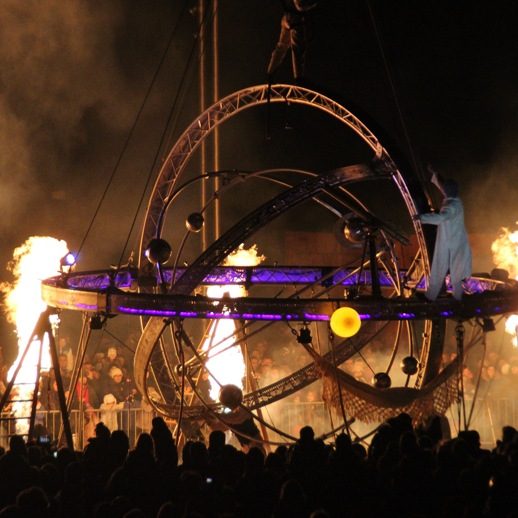 Galileo spectacle sous grue Artiste manipulant une structure lumineuse entourée de flammes, avec un public en arrière-plan.